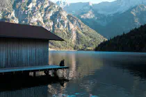 A person sitting at the edge of a dock in a mountainous lake